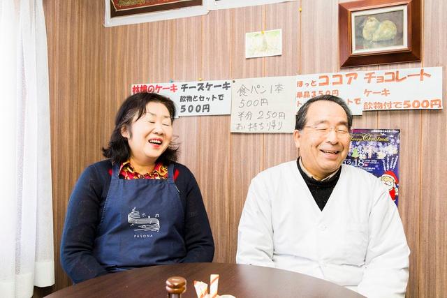 茶色の壁の前で椅子に座り微笑む長谷川さん夫婦の写真